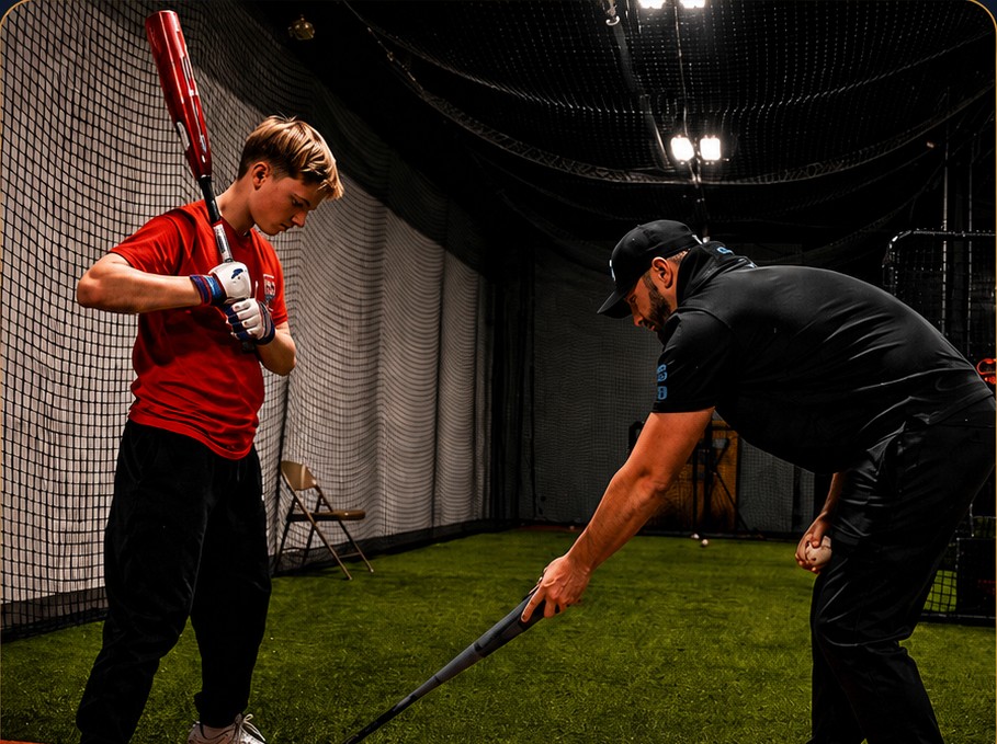 A baseball coach helping a young player with his batting stance inside an indoor batting cage. image.jpg